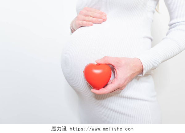 白色背景墙的一个美丽孕妇拿着红心在阳光明媚的夏日，怀孕、分娩、家庭观念、孕妇和户外心脏符号